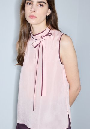Kobieta z długimi brązowymi włosami, ubrana w bluzkę bez rękawów w blady róż z ciemnoróżową lamówką i detalem z kokardką, stojąca na jednolitym jasnym tle.