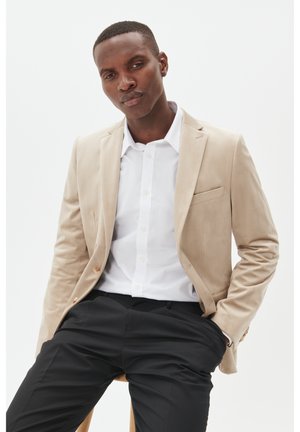 Beige blazer in soft fabric, single-button closure, notch lapels, breast pocket; paired with a white button-up shirt and black pants.
