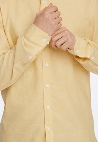  Lichtgele blouse met lange mouwen van een gestructureerde stof. Voorzien van witte knopen en verstelbare manchetten. Geknoopt langs de voorzijde.