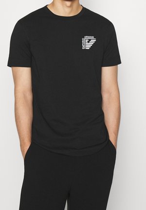 Mężczyzna w czarnej koszulce Emporio Armani z białym logo na klatce piersiowej, z rękami w kieszeniach, pokazany od szyi do bioder.