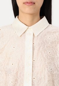 Light cream blouse featuring a floral embroidered design, with a collared neckline and button front closure. Soft, textured fabric.