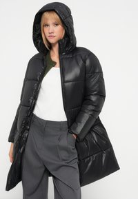 Casaco puffer preto com capuz, apresentando um design acolchoado e textura brilhante. Usado sobre uma blusa branca e calças cinzas.