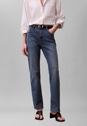 Personne portant un jean bleu taille haute, une chemise blanche boutonnée avec les manches retroussées, une ceinture noire et des sandales noires à talons blocs ouvertes à l'avant.