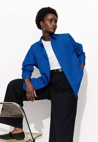 Femme aux cheveux bouclés naturels appuyée sur une chaise, portant une chemise bleu boutonnière, un tee-shirt blanc, un pantalon large noir et des mocassins noirs à clous.