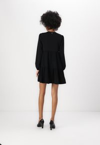 Robe noire courte avec des manches longues, design à volants et texture plissée. Fini avec un col rond et des boutons le long du dos.