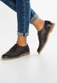 Scarpe oxford in pelle marrone scuro con dettagli perforati, suola chiara a contrasto e lacci, indossate con jeans in denim arrotolati.