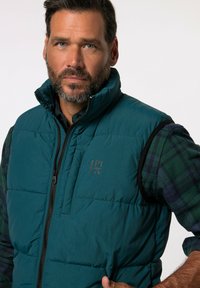 Teal puffer gilet met een rits aan de voorkant, met een borstzak en een logo. Draagt over een groen en marineblauw geblokt shirt. Gelaagd afwerking.