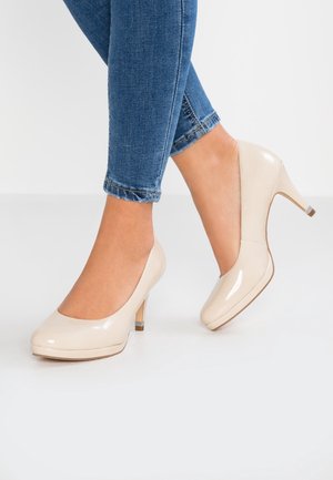 Kvinde iført blå skinny jeans og beige stiletter i laklæder, den ene fod fladt på jorden og den anden løftet på tæerne.