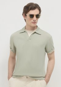 Polo shirt verde chiaro a maniche corte con scollatura a V e polsini arrotolati, realizzato in tessuto morbido. Indossato sopra una t-shirt color crema, abbinato a occhiali da sole.