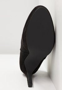 Černá semišová lodička na vysokém podpatku s špičatou špičkou a texturovanou černou podrážkou. Design se vyznačuje hladkým povrchem a minimálními akcenty.