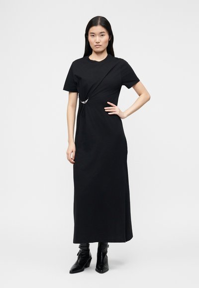 Γυναίκα με μακριά μαύρα μαλλιά που φοράει μαύρο midi φόρεμα με κοντά μανίκια, με λεπτομέρεια ρυτιδώματος στο πλάι και μαύρα μποτάκια μέχρι τον αστράγαλο, στέκεται με το χέρι στη μέση.