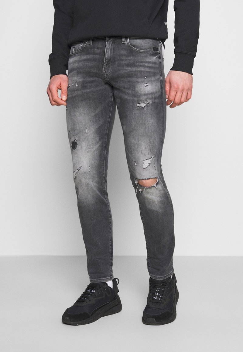 Man in slim fit, vervaagde zwarte ripped jeans met gaten en zwarte sneakers, staande tegen een neutrale achtergrond.
