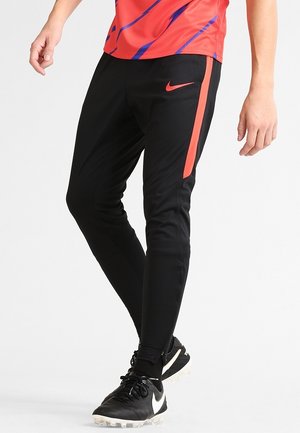 Mężczyzna noszący czarne i czerwone sportowe spodnie Nike oraz czarne korki, stojący na białym tle.