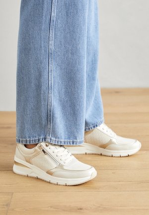 Crèmekleurige sneakers met textuuraccenten en een ritsdetail, gecombineerd met lichtblauwe wijde jeans, op een houten vloer.