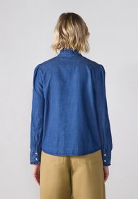 Denimskjorte med høy volangkrage, lange ermer med knappelukking og en lett innsnodd silhuett i mørkeblått stoff.