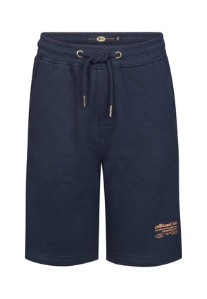 Jongen die een perzikkleurig T-shirt met coördinatietekst, marineblauwe korte broek, witte sokken en witte sneakers draagt, staand tegen een effen achtergrond.