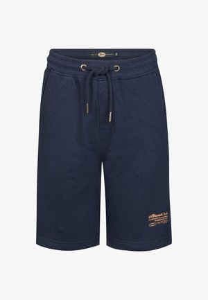 Jongen die een perzikkleurig T-shirt met coördinatietekst, marineblauwe korte broek, witte sokken en witte sneakers draagt, staand tegen een effen achtergrond.