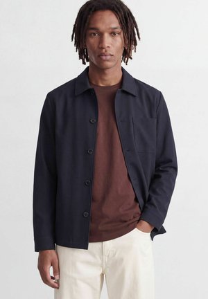 Mand med dreadlocks iført navy-blå skjortejakke over brun skjorte og hvide bukser, står mod en lys baggrund.