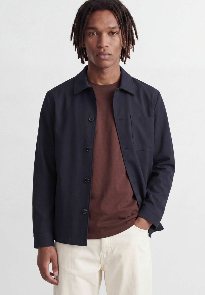 Homme avec des dreadlocks portant une veste bleu marine boutonnée sur une chemise marron et un pantalon blanc, debout devant un fond clair.
