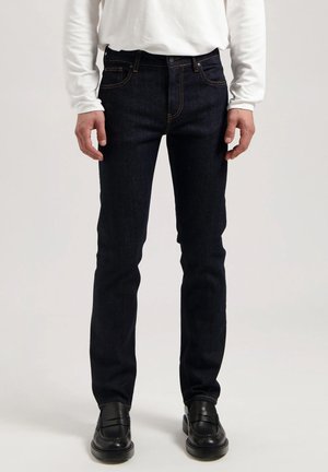 Man met donkerblauwe slim-fit jeans, wit overhemd met lange mouwen en zwarte leren loafers, staand tegen een effen achtergrond.