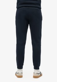 Marinefarbene Sweatpants mit taper Passform, elastischem Bund und Seitentaschen. Kombiniert mit weißen Sneakers, die schwarze Streifen und beige Akzente aufweisen.
