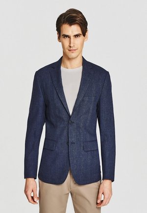 Mężczyzna ubrany w granatową marynarkę w kratę na jasnoszarej koszuli i beżowych spodniach, stoi na jednolitym jasnym tle.