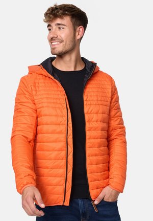 Orange polstret jakke med en sort hætteforing, lynlås lukning og tekstureret, quiltet design. Båret over en sort skjorte.