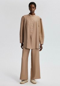 Tan, textured, oversized shirt with a high neckline and button details, paired with loose-fitting, matching trousers. Earthy tones.