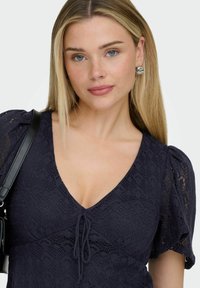 Jeune femme aux longs cheveux blonds portant des boucles d'oreilles argentées, une robe en dentelle noire à manches bouffantes et un sac à main noir, sur un fond uni.