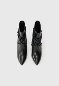 Pár čiernych členkových topánok s špicatou špičkou, s obtočenými remienkami a bočnými zipsami na jednofarebnom bielom pozadí.