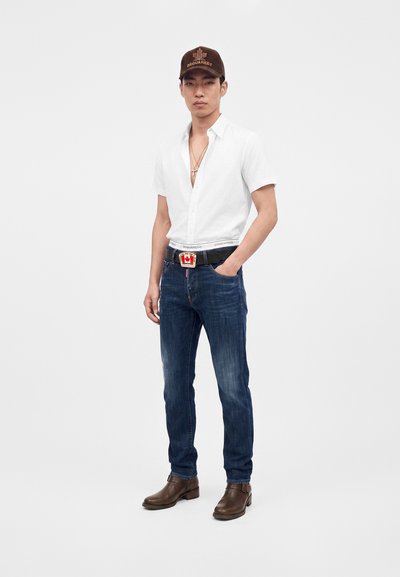 Jeune homme portant une casquette marron, une chemise blanche à manches courtes, un jean bleu avec une ceinture à boucle rouge et blanche, et des bottes marron.