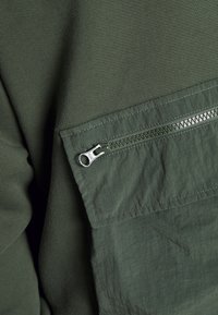 La prenda de color verde oliva presenta un tejido texturizado en un lado y una tela lisa y ligera con un bolsillo con cremallera en el otro.