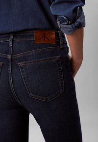 Blugi din denim albastru închis, cu design ajustat. Prezintă o etichetă din piele maro pe talie cu logo-ul "Calvin Klein" și buzunare standard în spate.
