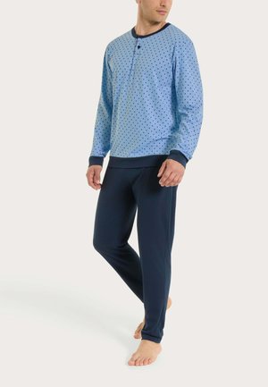 Uomo che indossa un pigiama a maniche lunghe azzurro chiaro con pois scuri e pantaloni del pigiama blu scuro, in piedi scalzo su uno sfondo semplice.