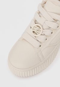 Baskette blanche en cuir lisse, avec un bout arrondi, une semelle en caoutchouc texturé et des lacets plats avec un accent circulaire sur le côté.