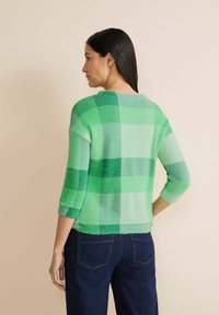 Zielony sweter w kratę z miękką, teksturowaną powierzchnią, rękawami o długości trzech czwartych oraz luźnym krojem. Połączony z ciemnoniebieskimi dżinsami.