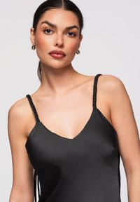 Femme aux cheveux foncés attachés, portant des boucles d'oreilles créoles dorées et un haut en satin noir à bretelles tressées, regardant la caméra avec assurance.