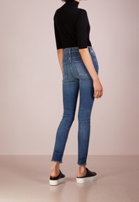 Jeans ajustados de color azul con un lavado desgastado, cintura alta y dobladillo deshilachado. Combinados con una blusa negra y zapatos negros sin cordones. Textura de denim suave.
