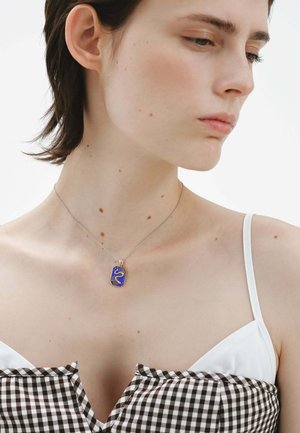 Collar de plata con un colgante rectangular que presenta un diseño azul y dorado. La modelo lleva una blusa de cuadros con tirantes blancos.
