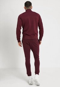 Bordowy dres z zamkiem przy kołnierzu. Tkanina jest gładka i elastyczna, z dopasowanymi rękawami i zwężanymi spodniami. Na nogach białe sneakersy.