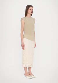 Top senza maniche beige con dettaglio legato sui lati, abbinato a una gonna midi in pizzo color crema con motivi circolari e scarpe bianche a slip-on con punta.