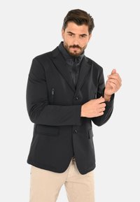 Giacca nera in tessuto soft-shell con chiusura frontale a bottoni, due tasche sul petto con cerniera e una texture liscia. Design aderente con colletto e maniche lunghe.