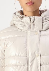 Detailní záběr osoby v lesklé béžové prošívané bundě s vysokým límcem zapnutým na zip i knoflíky a velkými zlatými kruhovými náušnicemi.