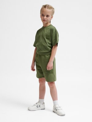 Dziecko z warkoczami stoi, ubrane w oliwkową koszulkę z krótkim rękawem i pasujące krótkie spodenki, białe skarpetki oraz białe trampki z szarymi akcentami.