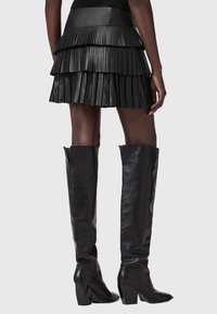 Μαύρη πλισέ μίνι φούστα με επίπεδη σχεδίαση, κατασκευασμένη από υφασμένο ύφασμα. Συνδυάζεται με ψηλές μαύρες δερμάτινες μπότες με τακούνι.