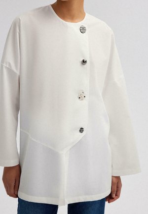 Blouse blanche asymétrique à manches longues avec quatre boutons distincts, portée avec un jean bleu, vue des épaules aux hanches.