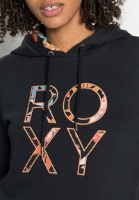 Mujer vestida con una sudadera negra con la palabra "ROXY" llena de coloridos patrones florales en el pecho, con la capucha forrada con una tela floral a juego.