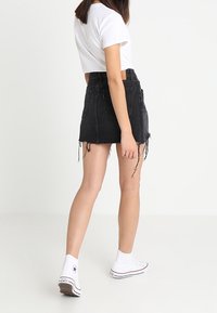 Kvinna som går bort ifrån kameran iklädd en vit t-shirt, svart fransig denimkjol och vita hög-top sneakers mot en enhetlig bakgrund.