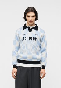 Νεαρός ενήλικας με κοντά σκούρα μαλλιά που φοράει πουλόβερ σε ανοιχτό μπλε και λευκό καρό με μαύρο γιακά και γκρι παντελόνι, στέκεται μπροστά σε λευκό φόντο.