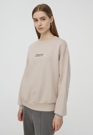 Felpa beige chiaro con scollo tondo, spalle scese, polsini a costine e un piccolo logo ricamato sul davanti.